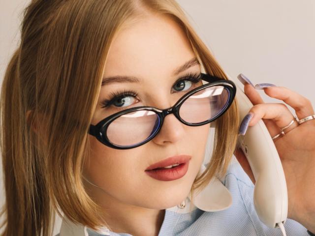 NicoleBrian with blonde pigtails, wearing black glasses, holding a white phone to her ear, has light skin, blue eyes, and purple nails, wearing a blue shirt.