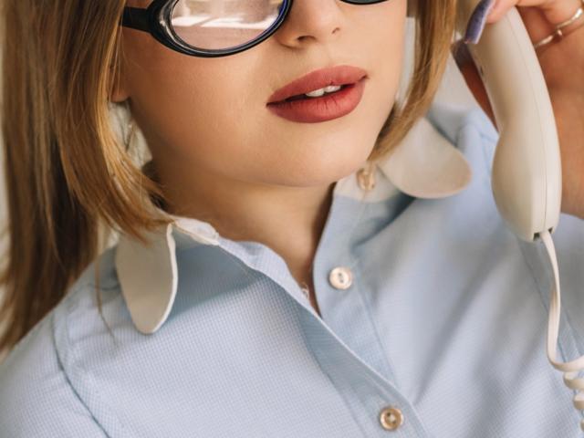 NicoleBrian with glasses, light blue shirt, white collar, holding a white phone to her ear, pink lips slightly open, blonde hair, soft skin, subtle makeup, close-up shot.