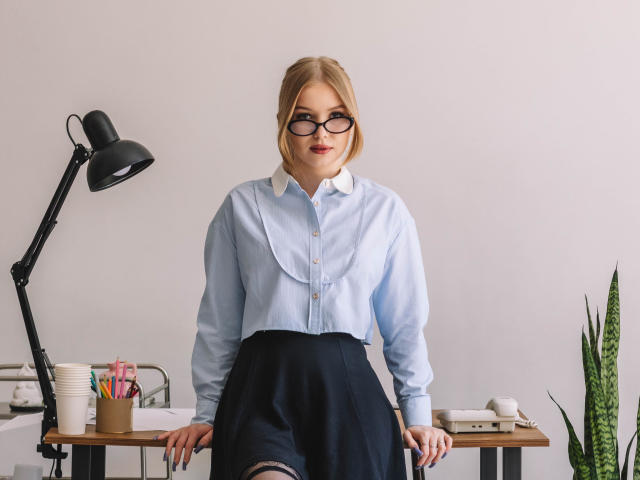 NicoleBrian with blonde hair, glasses, light blue shirt, black skirt, sitting at desk, hands on chair, black lamp, potted snake plant, pencils, white phone, and books.