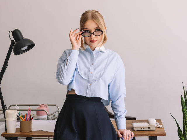 NicoleBrian with glasses, blue shirt, black skirt, touching nose, standing in front of desk with lamp, pens, and papers, looking at camera, blonde hair, cute, confident.