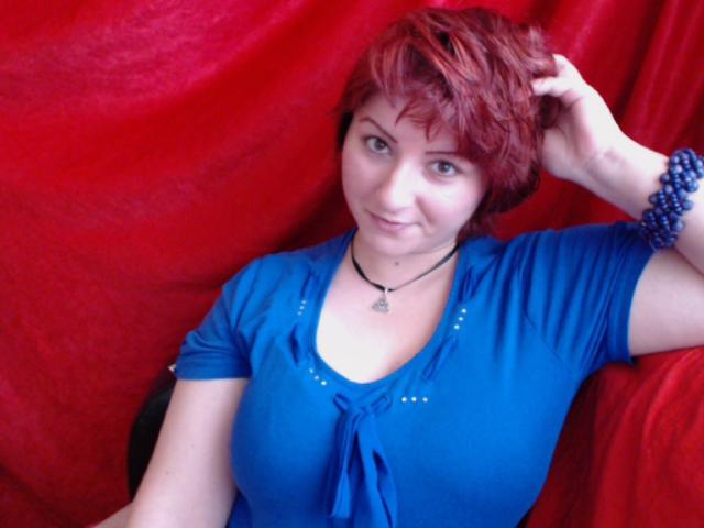PurrfectBoobs with red hair, wearing a blue top, has her hand in her hair, blue bead bracelet, necklace, smiling, sitting against a red fabric, showing her tits.