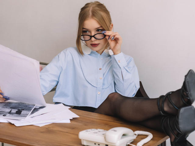 NicoleBrian, blonde, wearing glasses, blue shirt, black pantyhose, sitting at desk, legs up, black heels, white phone, papers, and laptop, looking at camera, slightly smiling.