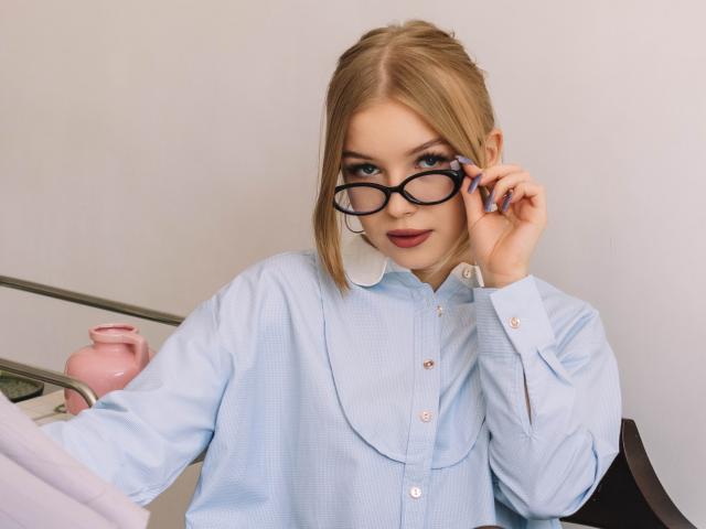 NicoleBrian with blonde hair, blue eyes, and glasses, wearing a light blue button-up shirt, adjusting her glasses, holding a pink teapot on a metal tray.