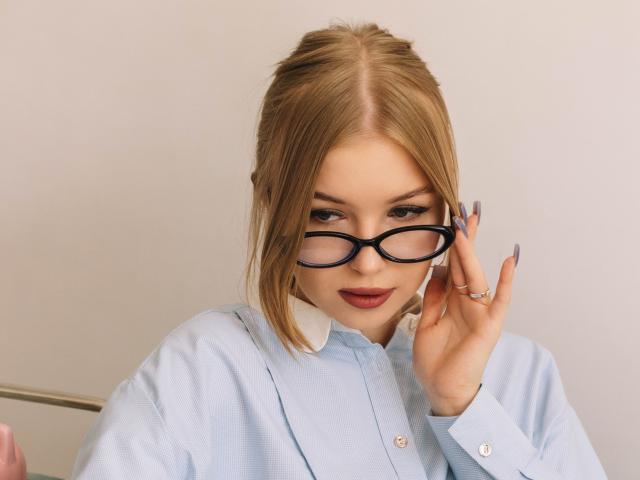 NicoleBrian with blonde hair, black glasses, and blue shirt, holding her glasses with long, manicured nails, looking down with a slight smile, showing her cute face and boobs.
