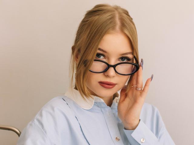 NicoleBrian with blonde hair, wearing glasses, light blue shirt with white collar, touching her face, has long nails, and a slight smile, showing her tits subtly.