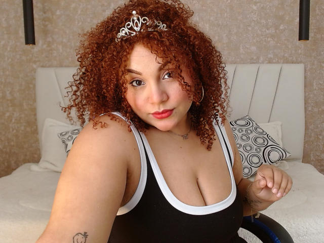 YeraCarey with curly red hair, wearing a silver tiara, black tank top, showing her tits, sitting in a wheelchair, red lipstick, tattoos on her arm and chest.