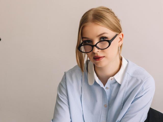 NicoleBrian has blonde hair in a braid, wearing black glasses and a light blue shirt with a white collar. Her ass is not visible. She looks at the camera.