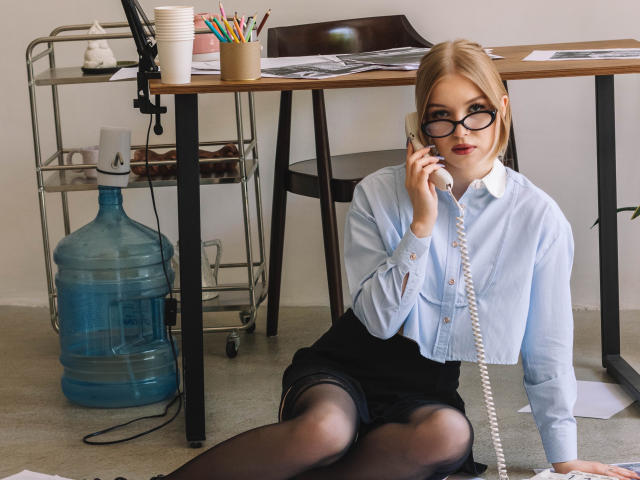 NicoleBrian with blonde hair, glasses, blue shirt, black skirt, and black stockings, sitting on carpet, talking on white phone, water cooler and desk in background.