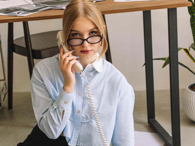 NicoleBrian with blonde hair, wearing black glasses, a light blue shirt, and black pants, sitting and talking on a white landline phone, smiling subtly.