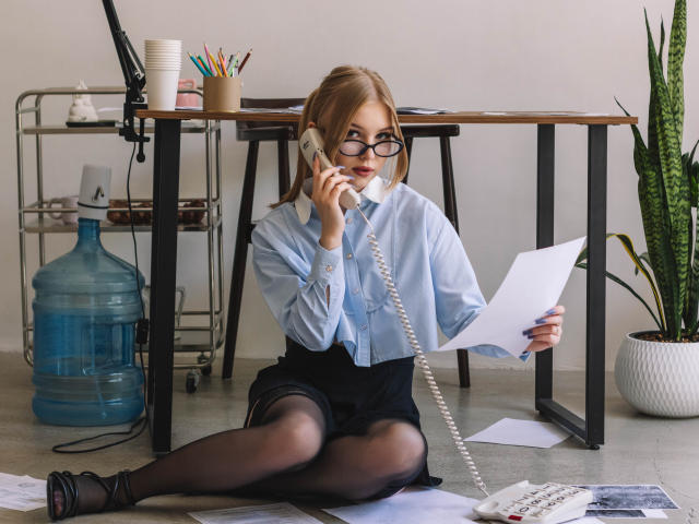 NicoleBrian with glasses, blonde, wearing a light blue shirt, black skirt, and black tights, sitting on the floor, talking on a white corded phone, holding papers.