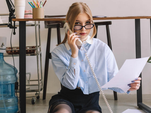 NicoleBrian, blonde, wearing glasses, light blue shirt, black skirt, holding phone to ear, holding papers, sitting on floor, water bottle, office desk, coiled phone cord, sexy.