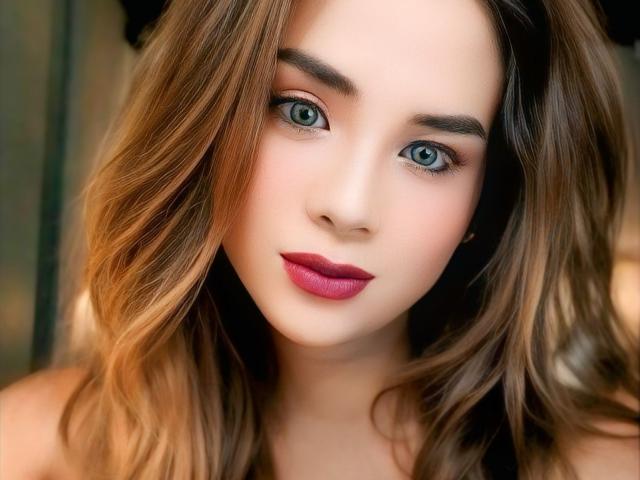 AlluringJhana has blue eyes, wavy brown hair, and pink lipstick, with a cute face and subtle makeup. Her tits aren't visible, just her pretty face.