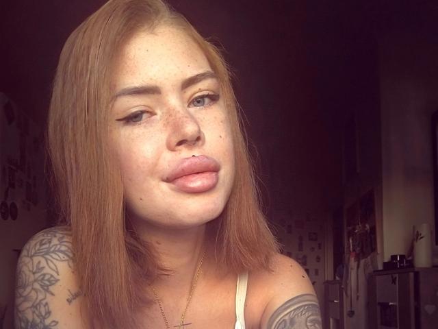 RoxyXXX – heiß, mit heller Haut, Sommersprossen und vollen, rosafarbenen Lippen – hat glattes, rotes Haar, Eyeliner im "Cat-Eye"-Stil und ein Tattoo an ihrer linken Schulter. Sie trägt ein weißes Tanktop..