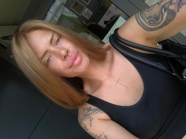 RoxyXXX - aantrekkelijk, met blond haar tot op de schouders, een zwarte tanktop, tatoeages op arm en borst, een neusring en een lichte glimlach, houdt een zwarte tas vast en toont een kleine kruistatoeage op de borst..