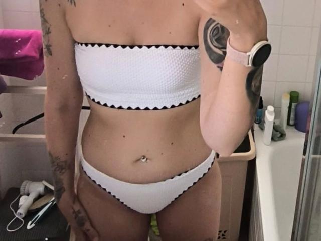 RoxyXXX, attraente in un bikini bianco senza spalline con bordi neri, mostra il seno, l'ombelico e i tatuaggi, tiene un telefono, indossa uno smartwatch, in un bagno..