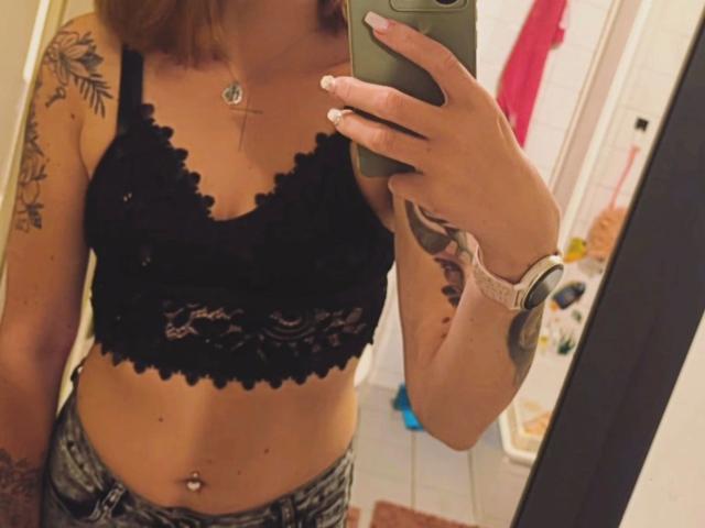 RoxyXXX-hot prend un selfie devant un miroir, vêtue d'un soutien-gorge en dentelle noire, montrant ses petites seins, son bras tatoué, son piercing au nombril, et porte un short en jean. Son téléphone est vert..