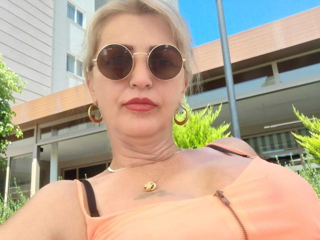 SexyCoco with blonde hair, wearing round sunglasses, gold hoop earrings, and a pink zip-up top, showing her boobs, and a small tattoo below her chest. Bright outdoor selfie.