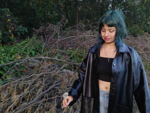 Aiiko with green-blue hair, black leather jacket, black crop top, and blue jeans, holding a small object, standing in front of tangled branches and greenery.