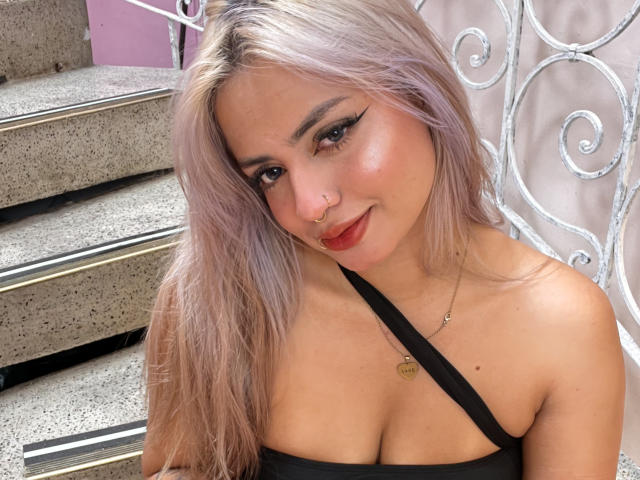 AlexaThoms, avec ses cheveux blonds platine, sa robe noire à bretelles, son anneau de nez et son eye-liner ailé, souriant à la caméra, montrant ses seins et son rouge à lèvres rouge..