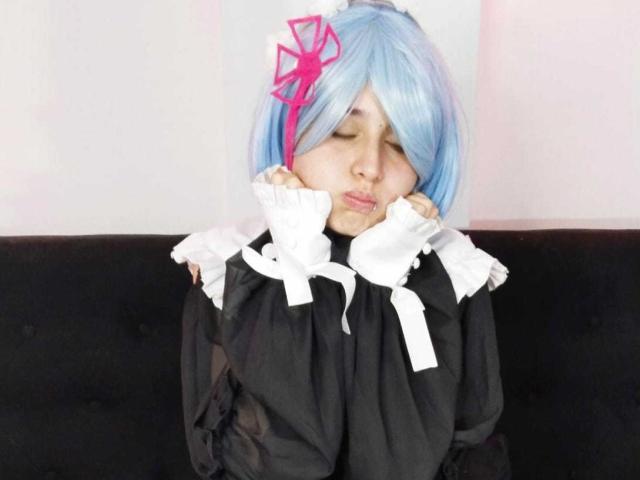 Aiiko with blue cosplay hair, pink bow, eyes closed, hands near face, wearing black and white outfit, sitting on black couch. She looks sleepy or bored.