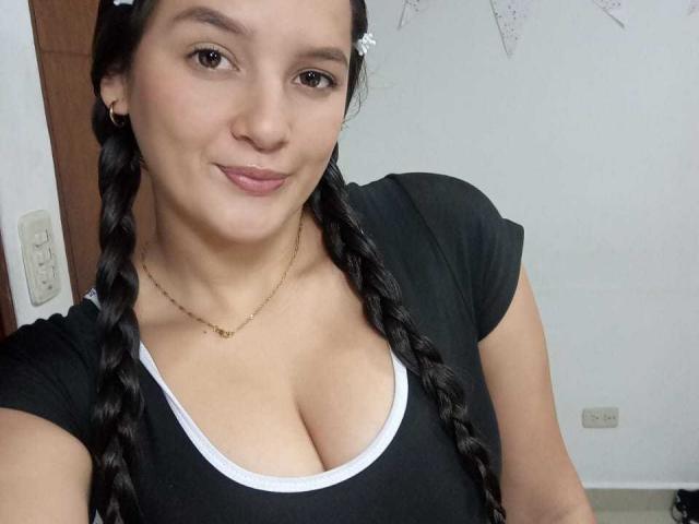 SamantaScott with braided black hair, light skin, brown eyes, and a small smile, wearing a black top with white trim, showing her big tits. She's wearing a gold necklace.