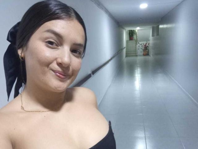SamantaScott with dark hair in a black bow, smiling, showing off her bare shoulders and small tits, wearing a gold necklace, in a shiny, tiled hallway.