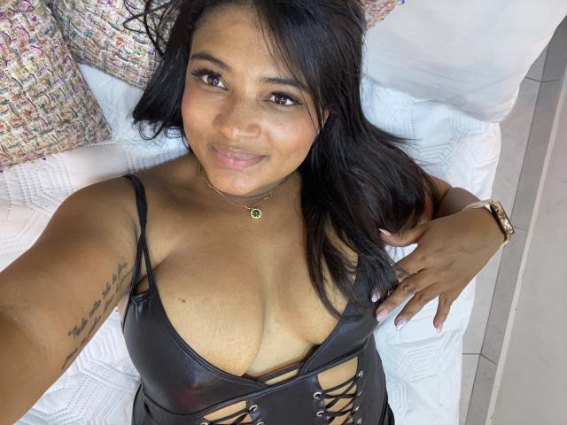 CameronJones with dark hair, tan skin, and a smile, lying on a bed, wearing a black leather top with cutouts, showing her large tits and a tattoo on her right arm.