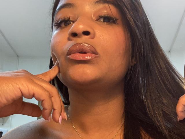 CameronJones has shiny brown lips, her finger touching her chin, with long dark hair and a gold necklace, brown eyes, and pinkish nails, in a slightly angled selfie.