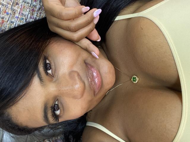 CameronJones with dark hair, brown skin, and pink glossy lips, wearing a yellow tank top, showing her tits, and a green gem necklace, resting her hand on her face.