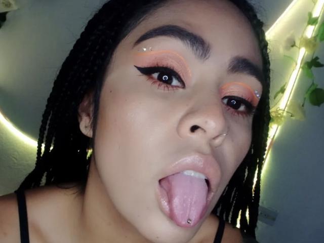 StellaBeckker's face with braided hair, pink and orange eyeshadow, black eyeliner, nose ring, tongue out, tongue piercing, and sparkling eye makeup. Her skin is light brown.