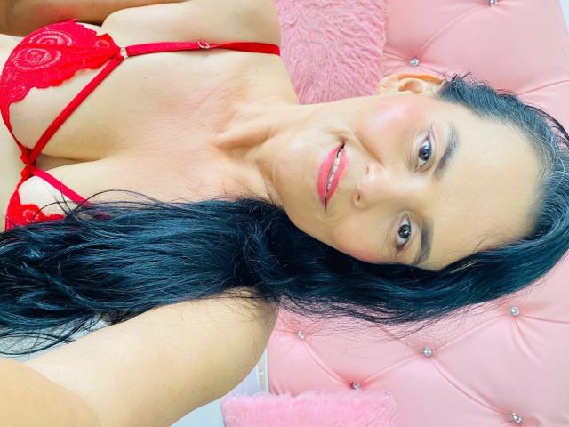 NoaLucia with black hair, red lace bra, and pink fluffy pillow, smiling at camera, showing her tits, red lipstick, and light skin, lying on pink tufted background.