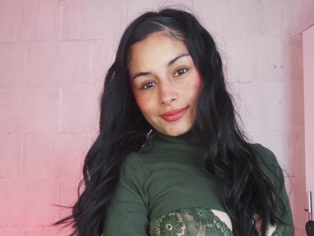 EmilyVv with long black hair, green sweater, and pinkish-red blush, smiling with red lipstick, nose ring, and brown eyes, standing against a white brick wall.