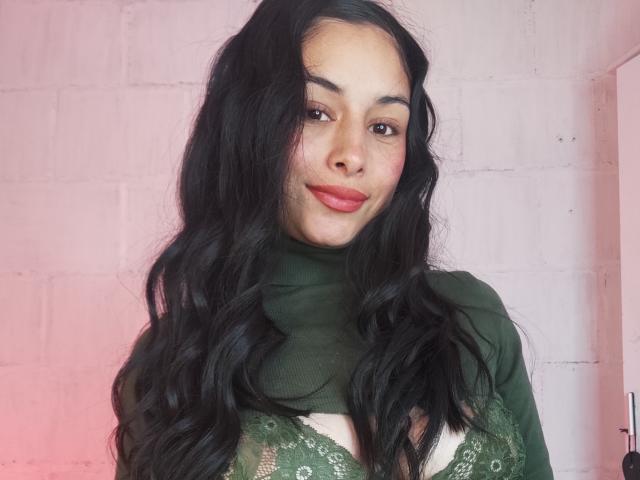 EmilyVv with long black wavy hair, green turtleneck, and green lace bra showing her tits, smiling with red lipstick, against a pinkish wall.