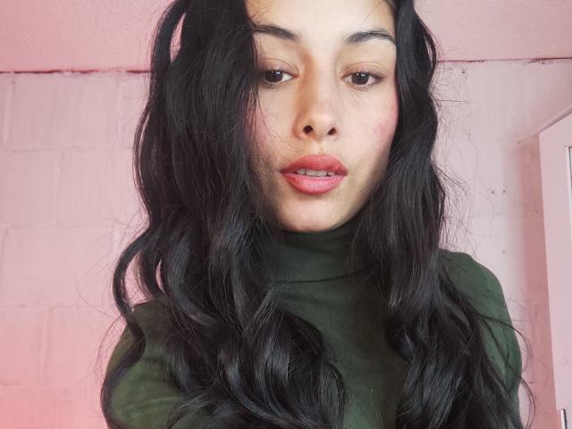 EmilyVv with long black wavy hair, green turtleneck, pinkish lips, light brown skin, and brown eyes, looking slightly to the side with a subtle pout.