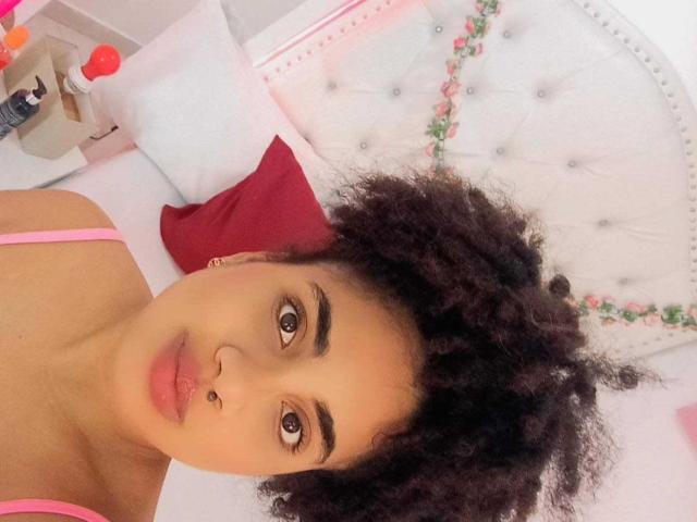 CuteAngelSwt with curly black hair, brown eyes, nose ring, pink top straps, lying on a bed with white and pink pillows, red pillow behind her head.