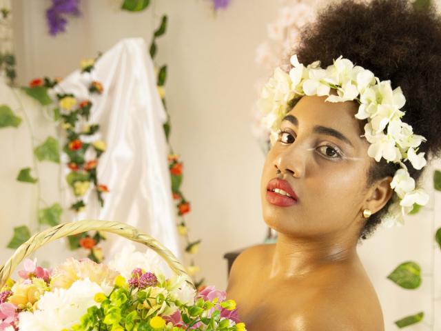 CuteAngelSwt with dark curly hair, wearing a white flower crown, red lipstick, and no top, holding a colorful flower basket, looking up with a gentle smile.