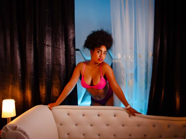 CuteAngelSwt with curly afro, wearing pink bra and purple panties, leaning on white tufted sofa, hands on top, blue and black curtains in background, soft lamp light.