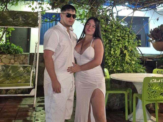 VickyAndDavid with large tits, wearing a white dress with a thigh slit, standing next to David in a white shirt and pants, both smiling in a garden.