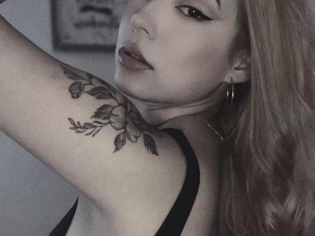 MissHayley with a tattoo on her arm, black eyeliner, and hoop earrings, looking over her shoulder, her blonde hair and lips slightly parted, wearing a black top.