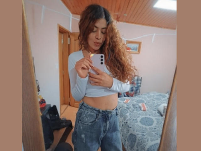 AlaiaLove, con cabello castaño rizado, vestida con una camiseta blanca de manga larga y pantalones vaqueros azules, tomando una foto de sí misma en un espejo, mostrando un abdomen plano y un trasero pequeño..