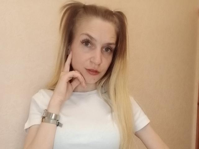 EmmaLili with blonde pigtails, white shirt, and silver wristwatch, touching her cheek with her finger, smiling subtly, showing her small tits and cute face.