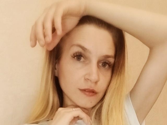 EmmaLili has blonde hair, light skin, and is looking at the camera with a slight smile. Her right arm is raised, touching her head, showing a ring on her finger.