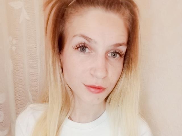 EmmaLili has blonde hair, blue eyes, and is wearing a white shirt. Her nose is small and her lips are slightly parted. She has long eyelashes and a subtle smile.