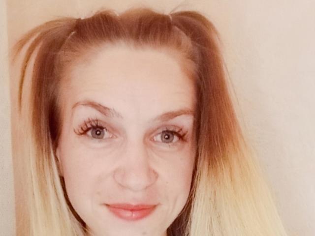 EmmaLili has blonde hair in pigtails, blue eyes, and a slight smile with red lips. She has long eyelashes and a fair skin tone. Her face is centered.
