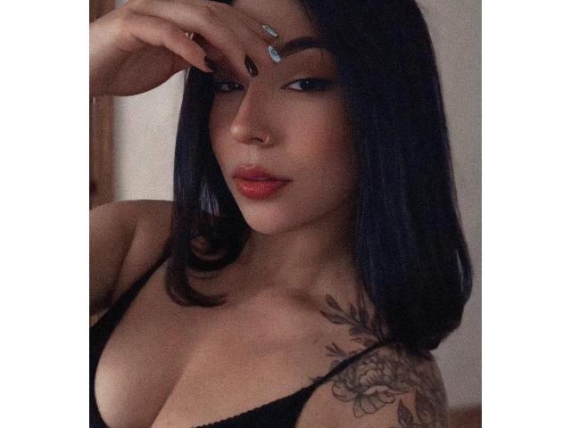 MissHayley with dark blue hair, black lingerie, nose ring, and tattoo on her right shoulder. She's touching her forehead with painted nails, showing red lips and a sultry look.