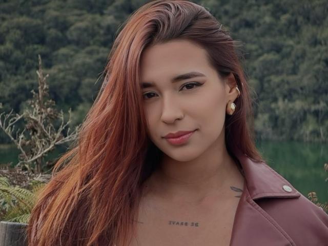 MissHayley with red hair, wearing a maroon leather jacket, small gold earrings, and a tattoo just below her tits. She's smiling subtly at the camera.