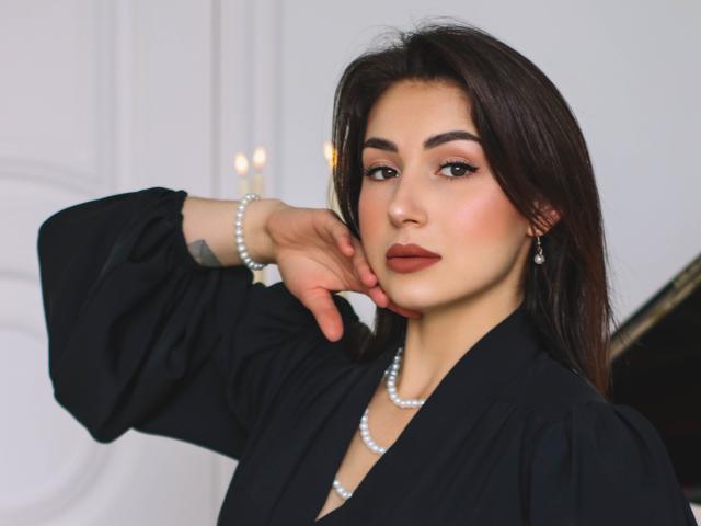 OliviaCluff with dark brown hair, wearing a black dress, pearl necklace, and bracelet, has a tattoo on her left wrist, and is touching her cheek with a hand.