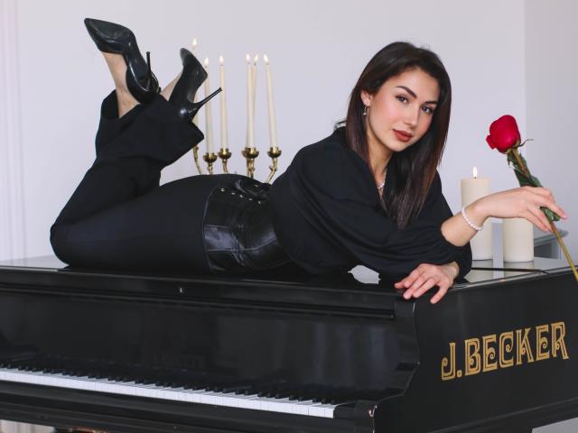 OliviaCluff lying on a black J. Becker piano, wearing black high heels and pants, with a rose in her right hand, candles in the background, and a sexy smile.