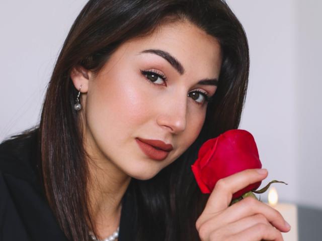 OliviaCluff with dark brown hair, holding a red rose, has subtle makeup, pearl earrings, and is wearing a black top, looking at the camera with a slight smile.