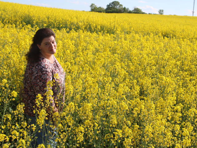 MollyBoobs-hot with curly brown hair, wearing a floral shirt, stands in a bright yellow wildflower field, looking back with a slight smile, under a clear blue sky.